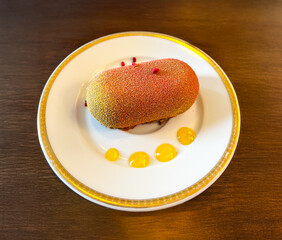 Wall Mural - Gourmet mousse dessert in orange and yellow glaze served on an elegant white plate with gold trim. Modern restaurant presentation on a wooden table.