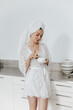 © vladdeep - Young caucasian mother with towel, on head in white sleeping robe, drink beverage from cup.