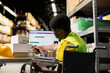 © DC Studio - Black wheelchair user in safety vest scanning barcode shipping labels, processing cargo orders on pc at desk. Warehouse clerk handles online shopping operations for retail large business.