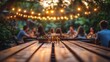 © sunny - Group of friends socializing outdoors at a wooden table with drinks and snacks under glowing string lights at dusk