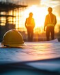 © oio - Yellow construction helmet resting on blueprints with two construction workers in reflective vests and helmets walking in the background during a golden sunset at a building site