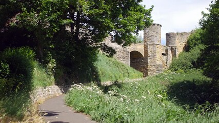Wall Mural - A medieval castle gate entrance in Scarborough, North Yorkshire, England, framed by tall trees and summer light. A majestic pathway bordered by stone walls and greenery.
