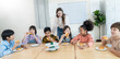 © paulaphoto - Happy group of diverse school children eating donuts together at desks with their teacher nearby, celebrating a fun classroom break.