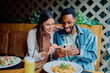 © we.bond.creations - Young couple enjoying a dinner date, smiling and laughing while looking at a smartphone together at a casual restaurant