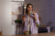 © Pixel-Shot - Young woman with glass of milk and cookie near open fridge in kitchen at night