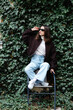 © nazariykarkhut - Stylish woman posing on a chair by a green ivy wall, wearing sunglasses and casual clothes