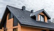 © Studicon - The image shows a newly constructed house featuring a gabled roof with dark gray metal tiles, two chimneys, and several windows, all set against a cloudy sky.