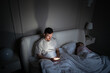 © DimaBerlin - Weary man awake scrolling smartphone late night unable to sleep in bedroom with wife. Insomnia issue, nighttime procrastination, digital distraction, sleep problem, mental restlessness, health concern