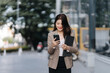 © Kritdanai - Smart Communication in the City: An elegant professional woman, immersed in her mobile phone, navigates the city with a digital device and documents, embodying contemporary urban life.