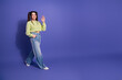 © deagreez - Young woman in trendy casual outfit smiling and waving hand while posing against a vibrant purple background for lifestyle shoots