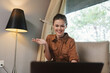 © SHOTPRIME STUDIO - Young woman in stylish brown shirt smiling while video chatting in a cozy room with modern decor, perfect for concepts like remote work and connection