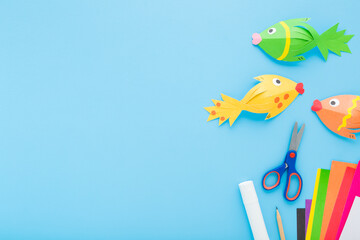 Wall Mural - Fishes, scissors, glue stick, pencil and colorful application paper on light blue table background. Pastel color. Closeup. Child created shapes. Empty place for text. Top down view.