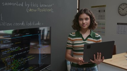 Medium portrait of smiling, confident Caucasian girl student holding laptop by blackboard with computer code, while presenting project on screen at programming class