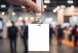 © Odesign - ID card mockup with lanyard, held by a person's hand. Lanyard and white card. In the background, out of focus, image of an event with stands