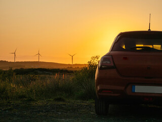 Wall Mural - A red car is parked in a field with a sunset in the background. The car is facing the sun, and the sky is orange and pink. Wind farm turbines in the background. Sightseeing modern technology concept