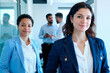 © pressmaster - Portrait of young adult Caucasian woman and young adult Black woman standing in modern office, smiling at camera while two young adult men having conversation in background