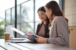 © wattana - Two professional women reviewing paperwork and working on a tablet in a bright modern office.