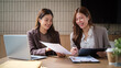© wattana - Two smiling businesswomen discussing paperwork and digital reports at a desk.