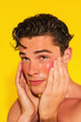 © Marut - Young man with wet hair and glowing skin gently applying facial oil, close up on face, yellow background, healthy skincare routine, fresh and confident expression