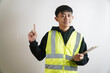 © yashis007 - Confident young Asian professional in safety vest and clipboard, pointing upwards, offers important guidance for workplace safety and operational efficiency