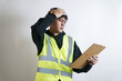 © yashis007 - A confused young male worker in a high-visibility vest holds his head in frustration while looking at a clipboard, feeling stressed by a mistake at work