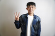 © yashis007 - Positive young Asian man with a genuine smile, confidently presenting three fingers as a clear visual count or a specific point in a sequence against a clean studio backdrop