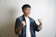 © yashis007 - Excited young Asian man in a denim jacket celebrating success with clenched fists, shouting with joy against a white studio background