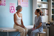 © DragonImages - Middle aged Caucasian woman with headscarf sitting on examination table, talking to young adult female healthcare worker wearing scrubs and gloves, in medical clinic cancer awareness posters visible