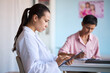 © DragonImages - Young adult Caucasian woman doctor using digital tablet while middle aged Caucasian woman patient filling medical form at desk in clinical setting, both focused on cancer consultation