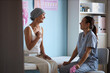 © DragonImages - Middle aged Caucasian woman with headscarf sitting on examination table, smiling and talking with young adult female nurse, in medical clinic with breast cancer awareness posters visible
