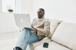 © SHOTPRIME STUDIO - A young Black man in a cozy beige sweater working on his laptop at home, radiating focus and comfort in a bright, minimalist setting