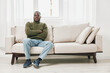 © SHOTPRIME STUDIO - A relaxed African American man in a cozy green sweater sits comfortably on a beige sofa, expressing calmness and confidence in a bright, minimalistic room