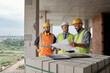 © pressmaster - Three workers wearing safety helmets and vests standing at construction site discussing building plans, one man holding blueprint while others listening attentively