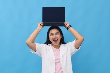 Very excited young Asian woman is lifting and showing blank laptop screen above her head, excited and cheerful, looking at camera, standing isolated on blue background.