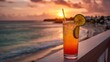 © Formoney - A glass of cocktail with a lime slice on top is sitting on a railing overlooking the ocean. The drink is a mix of orange and pink, and the sun is setting in the background. The scene is serene
