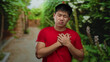 © Krakenimages.com - Young man in red shirt holding chest in park setting with green lush background, displaying emotion and introspection outdoor.