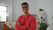 © Krakenimages.com - Young man in a red sweater and glasses smiling confidently with crossed arms in a bright indoor room featuring cozy decor and plants.
