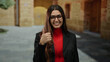 © Krakenimages.com - Hispanic woman smiling and giving a thumbs-up gesture outside at an old university campus wearing a red shirt and glasses.