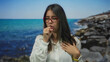 © Krakenimages.com - Woman coughing with closed eyes and hand on chest at seaside, wearing glasses, surrounded by ocean and rocks, shows health concern while standing outdoors in daylight.