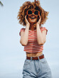 © SHOTPRIME STUDIO - Joyful woman with curly hair wearing a red striped crop top and high waisted jeans, posing outdoors under a clear blue sky with oversized sunglasses.
