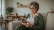 © NaesyaStudio - Elderly woman with grey hair wearing glasses and a green cardigan, sitting on a sofa and looking at her smartphone