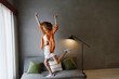 © SHOTPRIME STUDIO - Joyful young woman with curly hair in an orange shirt and white shorts dancing on a couch in a modern living room with minimalistic decor