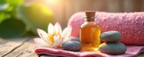 Essential spa items oil bottle, smooth stones, pink towel, lotus flower on wood. Natural setup creates serene wellness scene for therapy, relaxation. Warm sunlight enhances tranquil atmosphere.