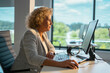 © Videophilia - Mature businesswoman focused on her work, sitting at a desk with a computer and headphones, processing data in a modern office with large windows overlooking a green landscape
