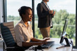 © Videophilia - Young businesswoman working on computer at her desk, concentrating on screen while male colleague stands observing the outdoor view from a large window in a bright office environment