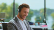 © Videophilia - Young man in smart-casual attire wearing a headset and microphone, smiling while providing customer service and support at a modern call center desk in a bright office