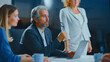 © Videophilia - Mature businessman leading a corporate meeting, actively discussing business strategy with colleagues around a table while holding eyeglasses and using a laptop in a modern office
