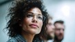 © Bussakon - A focused professional woman wearing glasses looks ahead in a bright office with blurred colleagues in the background