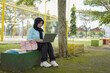 © Fajar - Young Muslim woman wearing a hijab diligently studying outdoors on a park bench with her laptop and books, embracing a modern lifestyle of remote learning and higher education