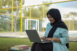 © Fajar - Young Muslim woman wearing a hijab diligently studying outdoors on a park bench with her laptop and books, embracing a modern lifestyle of remote learning and higher education